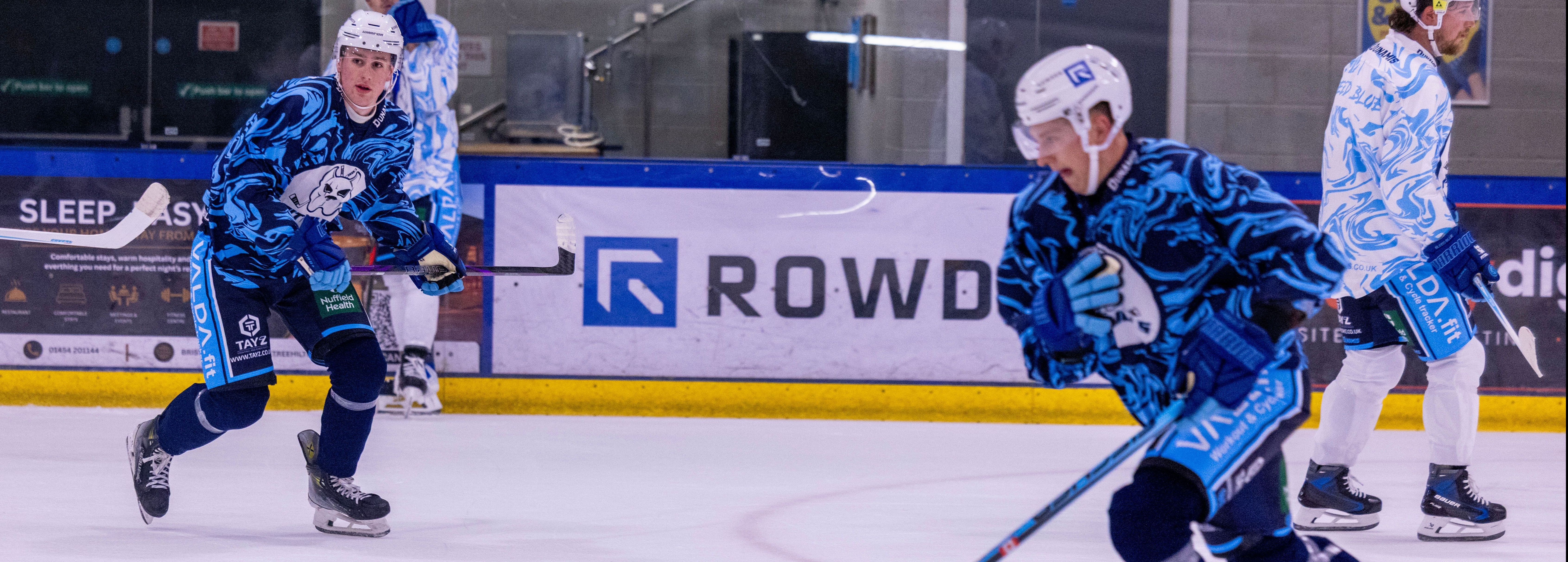 Two hockey players in blue patterned jerseys skate on an ice rink.